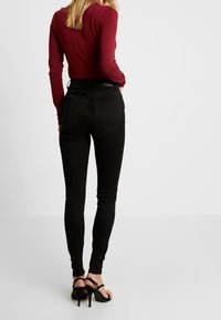 Woman wearing a fitted burgundy long-sleeve top, black skinny jeans, and black high-heeled sandals, facing away against a plain background.