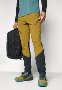 Gule og mørkegrå udendørsbukser med slim fit, der har en lynlås-lomme, parret med blå og grønne trail-sko og en sort rygsæk.