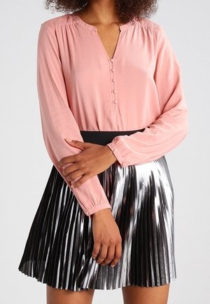 Vrouw draagt een roze blouse met lange mouwen en knopen en een geplooide metallic zilveren rok, poseert met gekruiste armen vooraan.