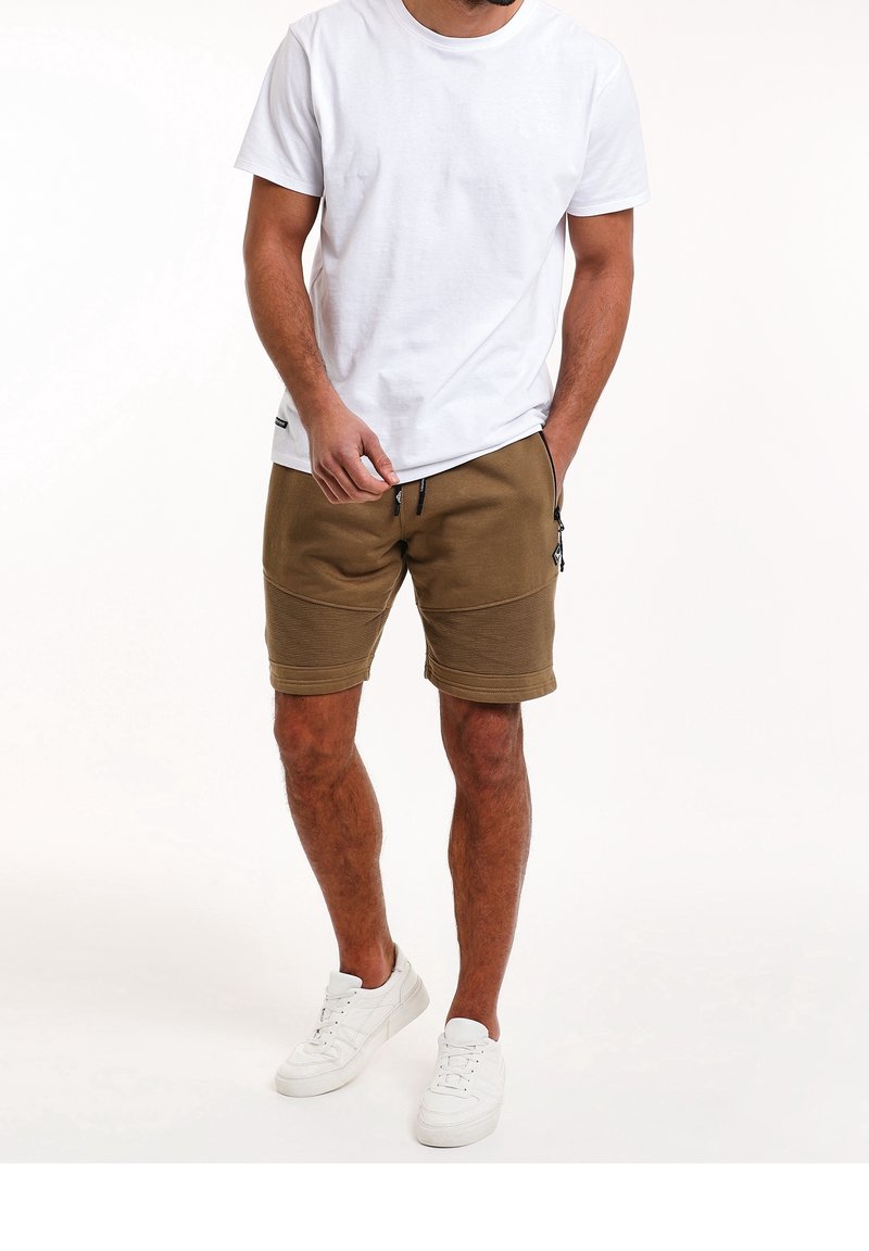 Homme portant un t-shirt blanc uni, un short marron jusqu'aux genoux avec des poches zippées, et des baskets blanches, debout devant un fond blanc.