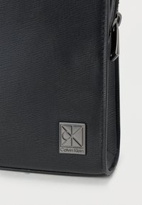 Μαύρη τσάντα με υφή, κατασκευασμένη από συνθετικό υλικό, με μεγάλο ασημένιο λογότυπο Calvin Klein και κλείσιμο με φερμουάρ. Μινιμαλιστικός σχεδιασμός.