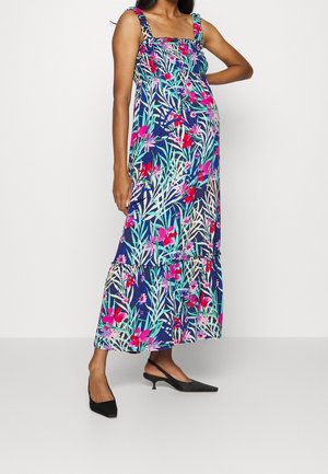 Pregnant woman wearing a blue floral sleeveless maxi dress with ruffle hem and black low-heeled slingback shoes, standing with hand on hip.