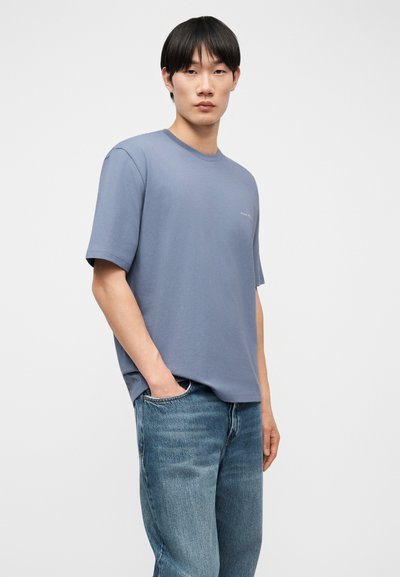 Νεαρός άνδρας στέκεται με το ένα χέρι στην τσέπη, φορώντας μια μονόχρωμη μπλε κοντομάνικη μπλούζα και μπλε τζιν, με φόντο λευκό.