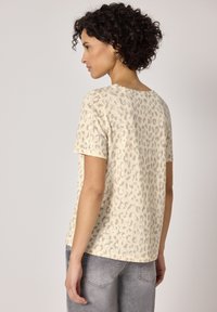 Naisharjainen nainen, jolla on vaaleanbeige lyhythihainen leopardikuvioinen paita ja haalistuneet harmaat farkut, sivuttain selin yksiväristä taustaa vasten.