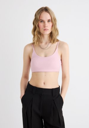 Tânără cu păr blond ondulat, până la umeri, purtând un top roz deschis scurt și pantaloni negri cu talie înaltă, pe un fundal simplu.