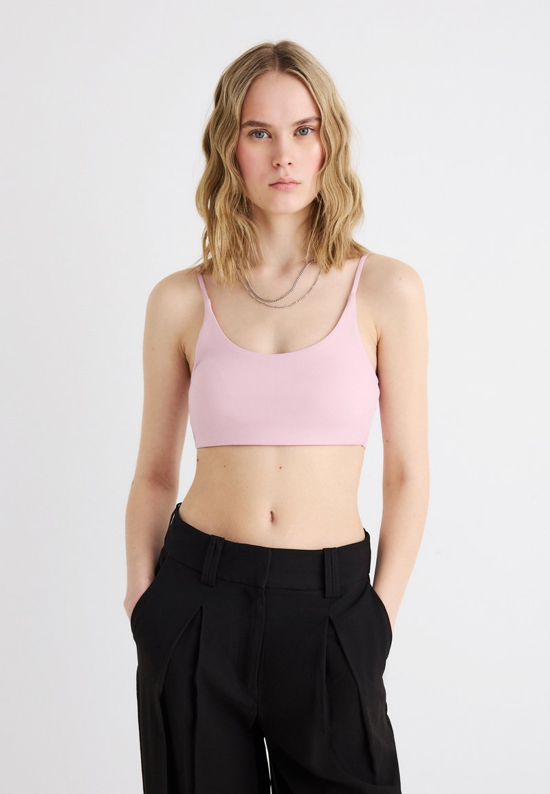 Νεαρή γυναίκα με ξανθά κυματιστά μαλλιά έως τους ώμους, που φοράει ένα ανοιχτό ροζ κοντό τοπ και ψηλόμεσα μαύρα παντελόνια, μπροστά σε απλό φόντο.