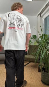 Balti marškinėliai su grafiniu dizainu, vaizduojančiu kavos ąsotį ir puodelį, užrašu „Originals Coffee Club“ ir „Adidas“ logotipu. Laisvo silueto. Juodos kelnės.