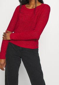 Roter gestrickter Pullover mit langen Ärmeln, gerippten Bündchen und Saum. Verfügt über eine strukturierte Oberfläche. Kombination mit schwarzen Hosen.