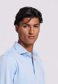 Lichtblauwe poloshirt gemaakt van zachte stof, met een knoopsluiting, een kraag en een gladde textuur, gedragen door een model dat naar voren kijkt.