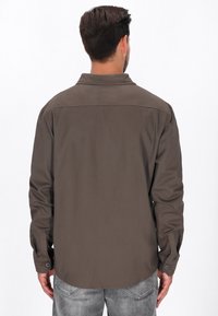 Bruine overhemd met knopen, kraag, lange mouwen en zichtbare stiksels. Gladde stof textuur, gedragen over lichtgrijze jeans.