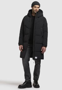 Sort polstret lang frakke med hætte, som har en lynlås fortil, sidelommer og et logo patch ved kanten, båret over mørke jeans og støvler.
