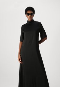 Rochie din satin negru cu guler înalt și mâneci scurte, având o croială lejeră și o textură fină. Modelul poartă ochelari de soare negri.