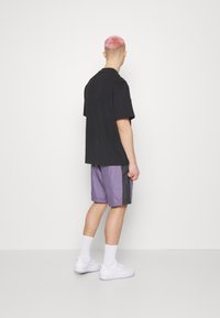 Černé oversized tričko, fialové šortky se šedými bočními panely, bílé tenisky, bílé ponožky po kotníky, viděno zezadu na jednobarevném pozadí.