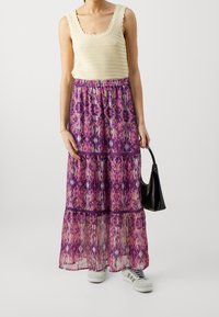 Débardeur tissé beige avec bords festonnés, associé à une jupe longue violette fluide ornée de motifs floraux colorés et d'un design en couches.