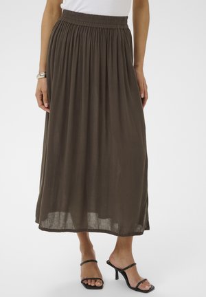 Femme portant une longue jupe plissée vert olive foncé, un haut blanc, un bracelet argenté et des sandales à talons ouvertes noires, debout devant un fond uni.