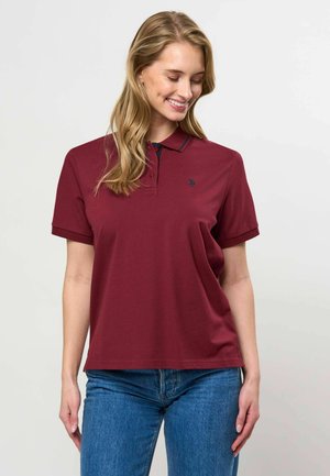 Jeune femme aux longs cheveux blonds portant un polo bordeaux à manches courtes et un jean bleu, souriante et regardant vers le bas.