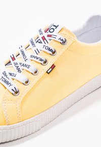 Yellow canvas sneaker with silver eyelets, white patterned laces, and a small logo tag. Textured white rubber sole with visible patterns.