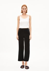 Witte mouwloze top met textuurdetails, gecombineerd met zwarte wijde broek. Model draagt donkere slip-on sandalen, tegen een effen achtergrond.
