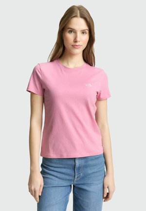 Młoda kobieta w gładkim różowym t-shircie z krótkim rękawem i niebieskich dżinsach, stojąca na neutralnym tle.