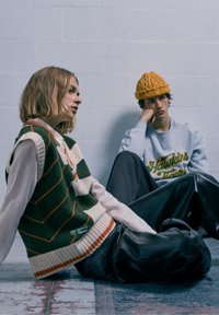 Strickweste mit grünen und cremefarbenen Streifen, getragen über einem weißen Hemd. Schwarze, glänzende Lederhose. Model mit einer gelben Strickmütze und einem grauen Sweatshirt.