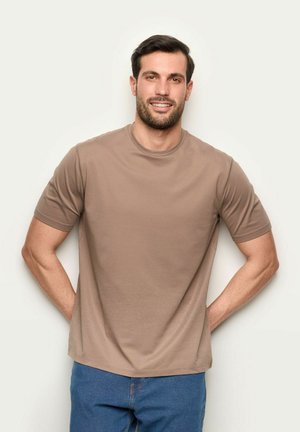 Uomo sorridente con capelli scuri e barba, che indossa una t-shirt beige e jeans blu, in piedi con le mani dietro la schiena su sfondo bianco.