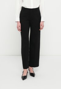 Pantalones negros de pierna ancha realizados en una tela suave, con cintura alta y líneas limpias, combinados con zapatos negros puntiagudos y una camisa blanca.