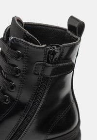 Detailní záběr na černou lesklou koženou botu se zipem na boku, černými tkaničkami, šitými detaily a látkovým poutkem na bílém pozadí.