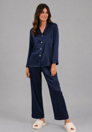 Femme debout portant un pyjama en satin bleu marine à manches longues boutonné et des chaussons blancs ouverts aux orteils et pelucheux sur fond gris.