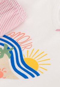 Tessuto di cotone bianco con un vivace design grafico che presenta palme, sole e onde. Include bordi di tessuto a strisce rosa e bianche.