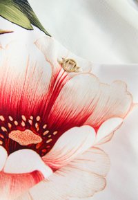 Tissu à motif floral avec une grande fleur rouge et des pétales blancs ; présente un petit logo doré attaché avec un cordon blanc.