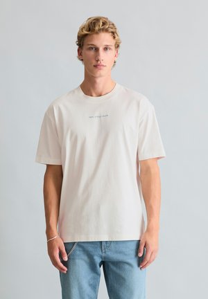 Młody mężczyzna z kręconymi blond włosami, ubrany w gładką, kremową koszulkę i jasnoniebieskie dżinsy na neutralnym tle.