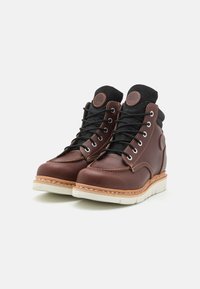 Botas de pele castanha com acentos em tecido preto, bico redondo, atacadores proeminentes, sola branca e detalhes de costura em contraste.