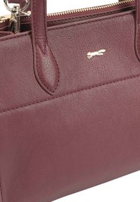 Bordeaux lederen handtas met een gestructureerde afwerking, uitgerust met twee handvatten en een gouden logo-accent. Ritssluiting aan de bovenkant.