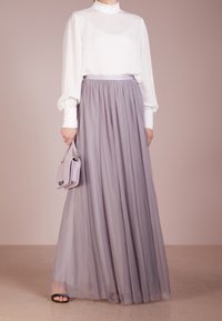 Femme portant un blouse blanche à manches longues, une jupe maxi en tulle légère de couleur lilas, des chaussures ouvertes noires et tenant un petit sac à main assorti.