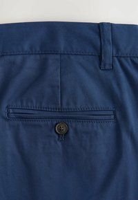 Poche arrière d’un pantalon bleu avec une poche passepoilée à bouton et une passants de ceinture au-dessus de la taille.