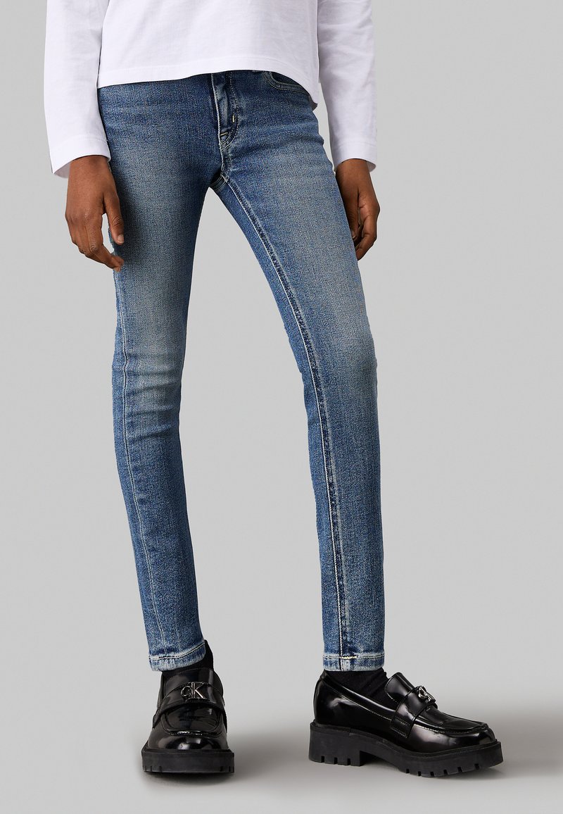 Schmale Jeans aus verblasstem, mittlerem blauen Denim mit leichter Abriebstelle, kombiniert mit schwarzen, glänzenden Slippern mit dicken Sohlen und einer dekorativen Schnalle.