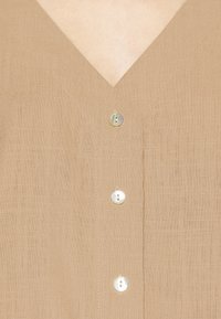 Top de lino beige con escote en V, que cuenta con tres botones blancos de concha. Textura ligera con detalles de costura visibles.