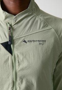 Veste vert clair en tissu texturé, dotée d'un col haut, d'une fermeture éclair à l'avant et d'un accent noir près de la poitrine avec un détail de logo.