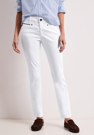 Kvinde iført tætsiddende hvide bukser, blå- og hvidstribet skjorte med opkørte ærmer og brune loafers, stående mod en ensfarvet baggrund.