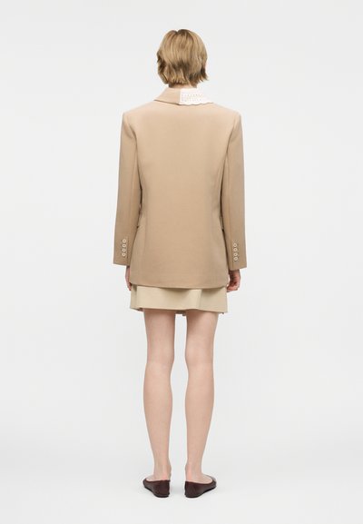 Άτομο με κοντά μαλλιά που φοράει μπεζ σακάκι, φούστα στο ίδιο χρώμα, λευκό πουκάμισο με γιακά και σκουρόκαφέ επίπεδα παπούτσια, με την πλάτη γυρισμένη σε λευκό φόντο.