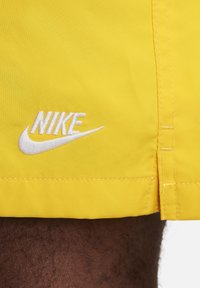 Shorts jaunes vifs en tissu lisse, avec un logo Nike brodé blanc sur l'ourlet et des détails de couture le long du côté.