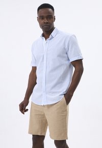 Camisa de manga corta abotonada en rayas azul claro y blanca, hecha de algodón, con cuello puntiagudo y un pequeño logo en el pecho, combinada con pantalones cortos beige.