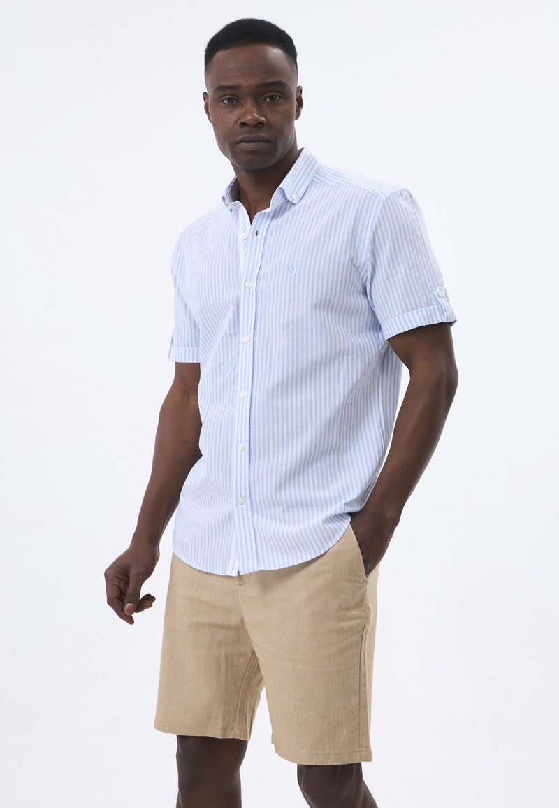 Camisa de manga corta abotonada en rayas azul claro y blanca, hecha de algodón, con cuello puntiagudo y un pequeño logo en el pecho, combinada con pantalones cortos beige.