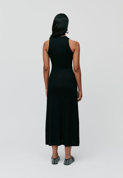 Black sleeveless maxi dress, form-fitting, with a smooth texture. Notable features include a clean back design and detailed shoes with studs.