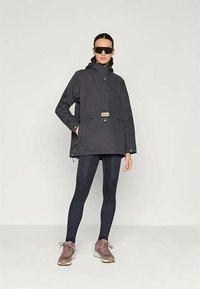 Μαύρη, αδιάβροχη ζακέτα με ψηλό λαιμό, μπροστινής τσέπης και ρυθμιζόμενο κορδόνι. Συνδυάζεται με σκούρες κολάν και καφέ αθλητικά παπούτσια.