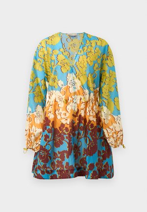 Vrouw staand in een kleurrijke bloemrijke jurk met opgeblazen lange mouwen, houdend een gevlochten zwart-witte mandtas, draagt bruine veterschoenen met platte zool.