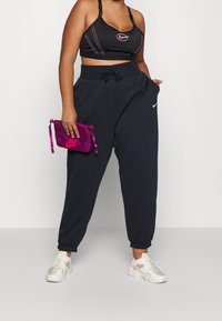 Kvinna i svart sport-bh och lösa svarta Nike sweatpants, som håller en liten lila Nike-påse, stående mot en neutral bakgrund.