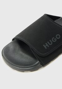 Svarte glidesandaler med strukturert såle, med en myk stoffoverdel og borrelåsrem. "HUGO"-logo trykket på siden.