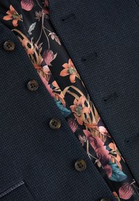 Marineblazer mit Struktur und zwei braunen Knöpfen; das Innenfutter zeigt ein florales Design in Rosa, Orange und Grün auf schwarzem Hintergrund.