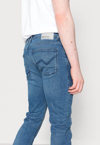 Person som bär blå denimjeans med en sydd bakfickedesign och en vit etikett märkt "DENIM TOM TAILOR," visad från sidan och bakifrån.